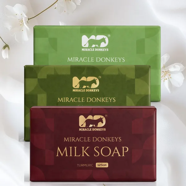 The three Miracle Donkeys milk soap packs in gold-foiled green and burgundy wrapping, resting on white silk with jasmine blossoms